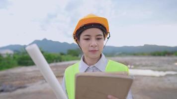 Portrait of young beautiful asian woman constructor, standing outdoor at outdoor construction site, looking at camera. Close up of female hard worker feel confident, wear safety vest and hard helmet video