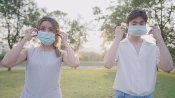 Young asian friends put on protective face masks from transmitted infectious diseases virus, standing outside in park new normal distancing, respiratory caring, thumps up gesturing, positive emotion video