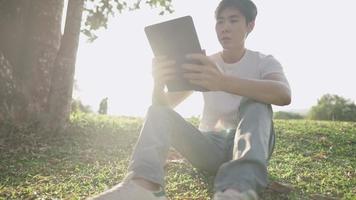 Young asian male using wireless tablet sitting relaxing at the park, on hot sunny good weather day, modern lifestyle with nature, message online dating application, portable gadget, browsing surfing