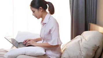 Aziatische werkloze vrouw zittend op de slaapkamer van een appartement met behulp van laptop op haar schoot met kussen, online op zoek naar een baan, werkkans, freelance werkende vrouw die thuis werkt met ochtendzon video