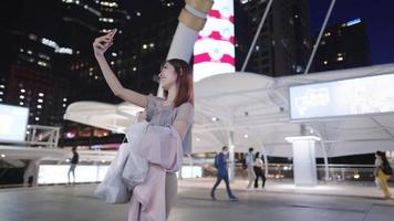 Attractive asian idol holding smartphone greeting to her online media followers while standing on city modern pedestrian in a nighttime, video calling or vlogging, digital marketing or online commerce