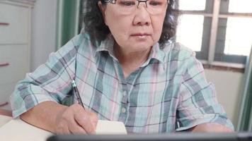 Aziatische senior vrouw draagt een bril, kijkt naar online lessen op een draadloze tablet, e-learning studie online, oudere generatie in de vrije tijd, schrijf op het notitieboekje, thuis, serieuze leerling video