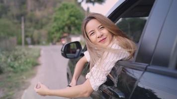 Happy blonde woman leaning out of car window while riding through country road, sticking her head out of moving auto and her long blonde hair blowing in wind, close up rear portrait and view video