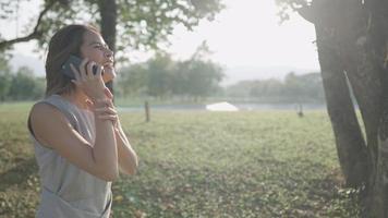 Fröhliches blondes Zopfhaar, das fröhlich telefoniert, während er tagsüber unter einem Schatten eines großen Baums im öffentlichen Park spazieren geht, süßes schüchternes Mädchen lacht, mobile Technologie für moderne Fernkommunikation video