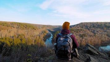 Young woman wearing colorful beanie and hoodie holding dog enjoys view of river and autumn forest under blue sky close back view slow motion video