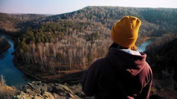 Brunette young woman in colorful beanie and hoodie enjoys hilly landscape view of river and forest under cloudless sky at sunset slow motion video