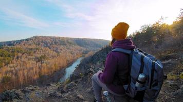 jeune femme en veste gonflée et bonnet avec thermos dans le sac à dos est assis sur le rocher du bord de la pente de la montagne en profitant de la vue sur la rivière et la forêt au ralenti video