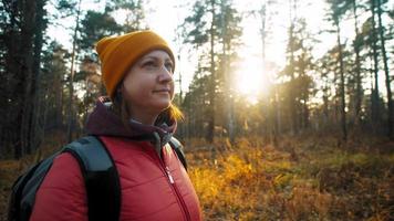 Brunette-haired woman in beanie and autumn jacket walks and enjoys coniferous forest view at bright orange sunset closeup slow motion video