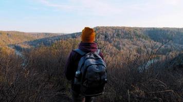 Young woman wearing hoodie and beanie with thermos in backpack strolls along mountain forestry slope against highland landscape slow motion