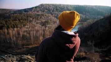 Young woman with brunette hair in beanie and hoodie enjoys curving river and forest view standing on hill top at sunset close backside slow motion video