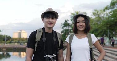 Side view of Happy asian traveler couple with hat smiling and looking at camera in the park. Joyful young blogger man and woman greeting with camera at park. Hobby and Lifestyle concept.