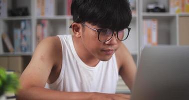 Closeup portrait of Focused freelancer man in singlet and eyeglasses works on laptop at home. Exhausted Asian young man with eyeglasses looks at computer notebook. Work from home and freelance concept video