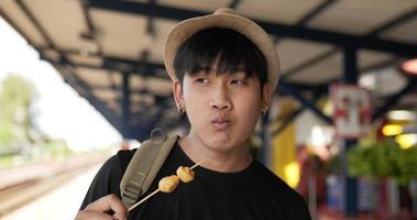 close-up de homem jovem viajante asiático comendo almôndega e olhando para a câmera na estação de trem. feliz macho faminto comendo aperitivo. conceito de transporte, viagens e comida.