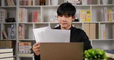 sério jovem empresário asiático verificando a correspondência de papelada corporativa sentado na mesa de escritório em casa. exausto empresário masculino lendo documentos, analisando papéis financeiros no local de trabalho.