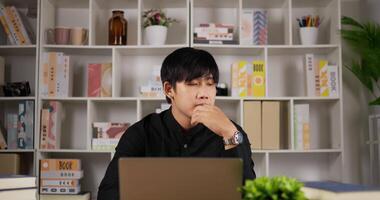 porträtt av asiatisk frilansman som arbetar framför laptop och tänker på hemmakontoret. allvarlig ung affärsman ser frustrerad tänker på arbete och sitter på arbetsplatsen. video