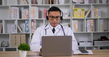 retrato de médico masculino asiático profissional de jaleco branco e fone de ouvido fazendo teleconferência no computador portátil na sala. consulta paciente a distância online por webcam. conceito de telemedicina. video