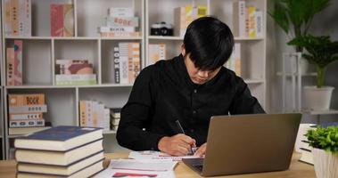 Portrait of Asian man hands writing information in strategy plan. Business man freelancer sits at table in front of laptop writes with pen on white paper in document plans analyzes in home office. video