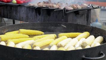 milho em água quente como comida de rua em uma feira ou festival de comida de rua. deliciosas espigas estão prontas para comer. milho amarelo em uma panela grande quente está sendo cozido. cozinhar legumes e carne para venda. video