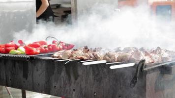 close-up van het braden van shish kebab op een spies en gegrilde groenten op een grote grill. kookproces. close-up zonnige buitenchef draait vlees op de grill. sappig varkensvlees of kip is rookgekookt. video