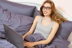 Happy smiling woman in glasses lying down the bed in front of her laptop in the morning bedroom photo