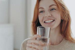 retrato de una feliz y saludable mujer pelirroja sosteniendo un vaso transparente de agua mineral pura
