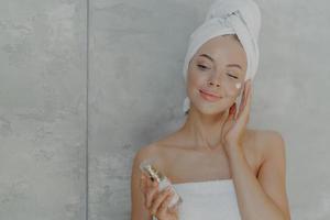encantadora mujer europea aplica humectante facial sostiene una botella de loción corporal, tiene una piel sana, tez bien arreglada usa una toalla envuelta en la cabeza después de ducharse, posa contra un fondo gris