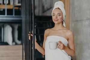 Photo of satisfied young female model wears cream on face, stands satisfied after taking bath, wrapped in white soft towel, drinks coffee or tea, poses over home interior. Anti aging treatment