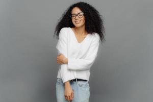 Indoor shot of sincere woman keeps hand partly crossed, dressed in white jumper and jeans, has eyes closed, enjoys happy moment in life, isolated over grey background. Positive emotions concept photo