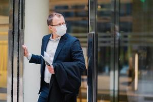 Respiratory protection, coronavirus, dangerous disease. Serious man stands in doors of office building, wears medical mask, holds modern cellphone, newspaper to read article about covid-19 treatment