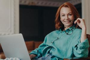 Indoor shot of pleasant looking redhead woman has good time at home, works on computer, wears casual shirt, holds eyeglasses, feels comfort, enjoys domestic atmosphere. Teacher teaches by video call photo