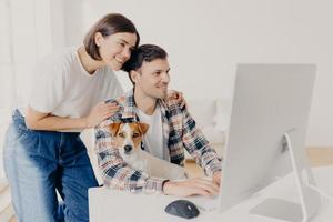 la imagen de una mujer toca suavemente los hombros de los maridos, se ve positivamente en el monitor de la computadora, navega por Internet, compra muebles en línea para su nuevo apartamento. hombre feliz trabaja en un dispositivo moderno con perro foto