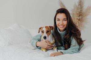 Photo of cheerful woman embraces dog with love, spend free time together, expresses tender feeling and emotions, falls in love with pet, lie on comfortable bed. Positive emotions, animals and care