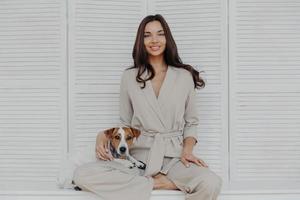 Horizontal shot of pretty relaxed woman dressed in beige costume, plays with favorite dog at home, being in love with domestic pet, expresses affection, being in good mood. Family, lifestyle concept photo