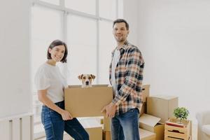 Happy family couple hold carton box with small puppy, stand indoor against big window, glad to become homeowners, unpack things in own apartment, looks gladfully at camera. Family moving day photo