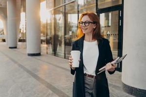 Female model holds touchpad notebook and cup of takeaway coffee poses outdoor during sunny day photo