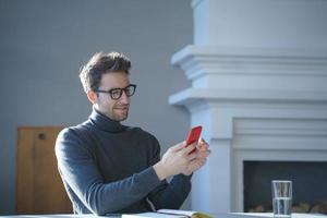 Young handsome german man in glasses sitting at table and using mobile phone at home photo