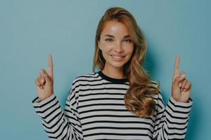 Photo of lovely smiling young woman points fingers up shows promo logo overhead