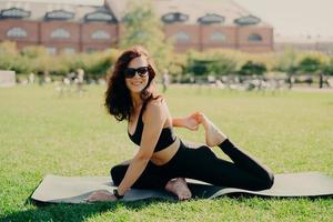 Happy fit young woman does stretching workout on fitness mat practices yoga outside dressed in activewear has strong body breathes fresh air outdoor leads active lifestyle. Sport fitness concept