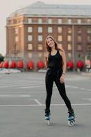 Full length shot of active smiling woman enjoys extreme sport rides on rollerblades dressed in black comfortable clothes stands outdoors learns rollerskating. People leisure recreation concept photo
