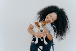 Glad dark skinned girl plays with jack russell terrier dog, have fun together, poses against white background, dressed casually. Happy Afro girl poses with pet indoor. People, animals, fun concept photo