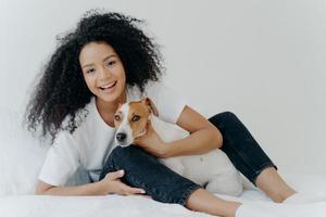 Horizontal shot of glad Afro woman rests in bed with dog, have playful mood, pose together in bedroom against white background. Girl relaxes at home with jack russell terrier. Sweet funny moment photo