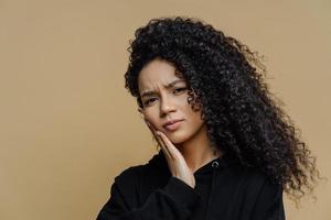 Headshot of displeased curly haired woman suffers from toothache, touches cheek, needs to see dentist, wears black sweatshirt, isolated on beige background. Tooth pain and dentristy concept.