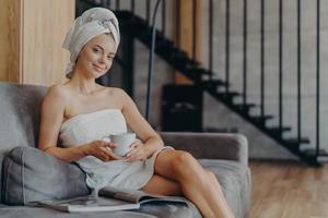 Horizontal shot of pleased young European woman smiles gently poses wrapped in towel drinks coffee poses on couch in living room reads magazine, applies beauty cream on face after taking shower photo