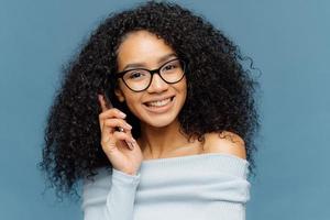 Portrait of satisfied African American woman talks on mobile phone, enjoys nice conversation, tells news to best friends, smiles broadly as hears funny story, isolated over blue background. Technology photo