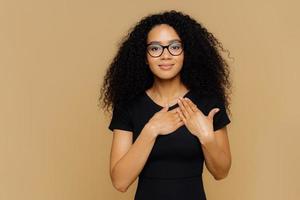 hermosa mujer de piel oscura mantiene las palmas cerca de escuchar, expresa gratitud, agradecimiento, tiene una expresión facial amistosa, usa anteojos y una camiseta negra casual, aislada sobre fondo marrón foto