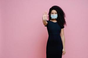 brote de covid 19, enfermedad viral. foto de una mujer étnica con el pelo rizado hace un gesto de parada con la palma, dice no al coronavirus, usa una máscara protectora estéril para evitar el virus, vestida de negro