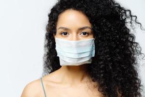 Serious Afro American woman tries to stop virus and epidemic disease, stays at home during infectious outbreak, wears medical mask, isolated on white background, being hospitalized, diagnosed photo