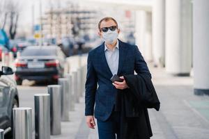 Businessman in black elegant clothes, medical mask, holds smartphone in hands, walks through busy city with many transport, protects himself from coronavirus during pandemic. Virus protection photo