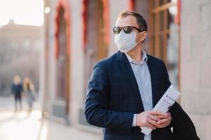 Working in pandemic situation. Business worker or entrepreneur wears medical mask for coronavirus protection stands beside office building outdoor holds newspapers concentrated somewhere into distance photo