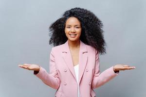 Isolated shot of cheerful female business worker with Afro hairstyle, wears elegant formal violet jacket, raises both palms, presents some product, smiles pleasantly, isolated over grey background. photo